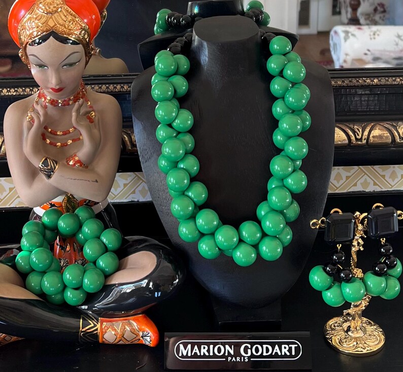 May include: A set of green and black beaded jewelry, including a necklace, bracelet, and earrings. The necklace and bracelet are made of large, round beads, while the earrings are smaller and have a dangling design. The jewelry is displayed on a black mannequin and a gold stand.