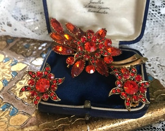 Puede incluir: Un conjunto de tres broches vintage de pedrería roja y dorada en una caja de terciopelo azul. El broche más grande es de diseño floral con una gran pedrería roja en el centro. Los broches más pequeños son en forma de estrella.