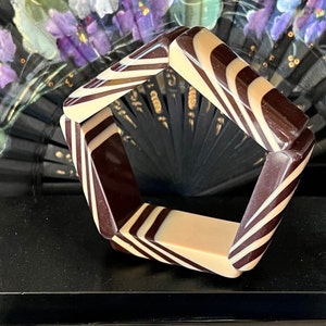 May include: A pentagonal bangle bracelet featuring a glossy, alternating pattern of dark brown and cream-colored stripes. The bracelet is displayed in front of a black base with the white text MARION GODART PARIS printed on the front surface.