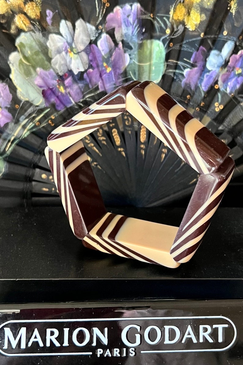 May include: A pentagonal bangle bracelet featuring a glossy, alternating pattern of dark brown and cream-colored stripes. The bracelet is displayed in front of a black base with the white text MARION GODART PARIS printed on the front surface.