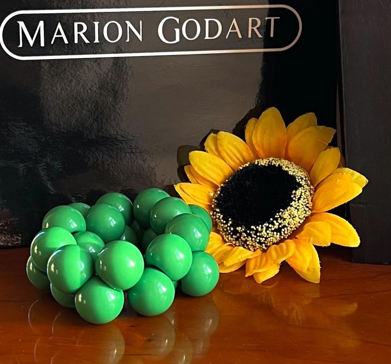 May include: A cluster of bright green round balls arranged in a circular shape, resembling a flower bud. A yellow sunflower with a dark brown center is in the background.