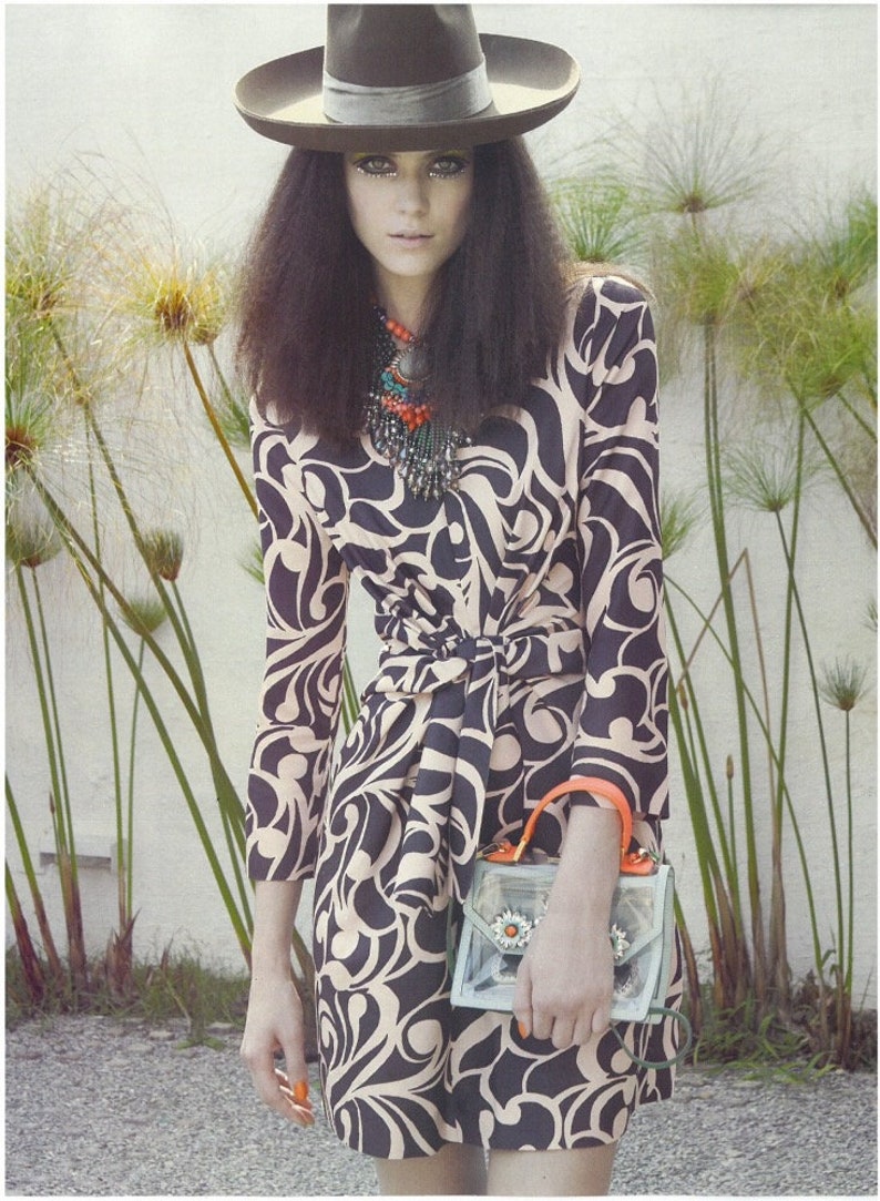 May include: A dark brown fedora hat, a multi-strand beaded necklace with orange and turquoise accents, and a knee-length dress featuring a black and beige swirling pattern. A clear handbag with orange handles and floral embellishments is held.