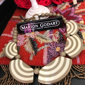 May include: A striking necklace with large, fan-shaped pendants, cream and taupe striped design. The necklace has round, taupe beads and a tag reading "MARION GODART PARIS". It is displayed on a red, embroidered surface.