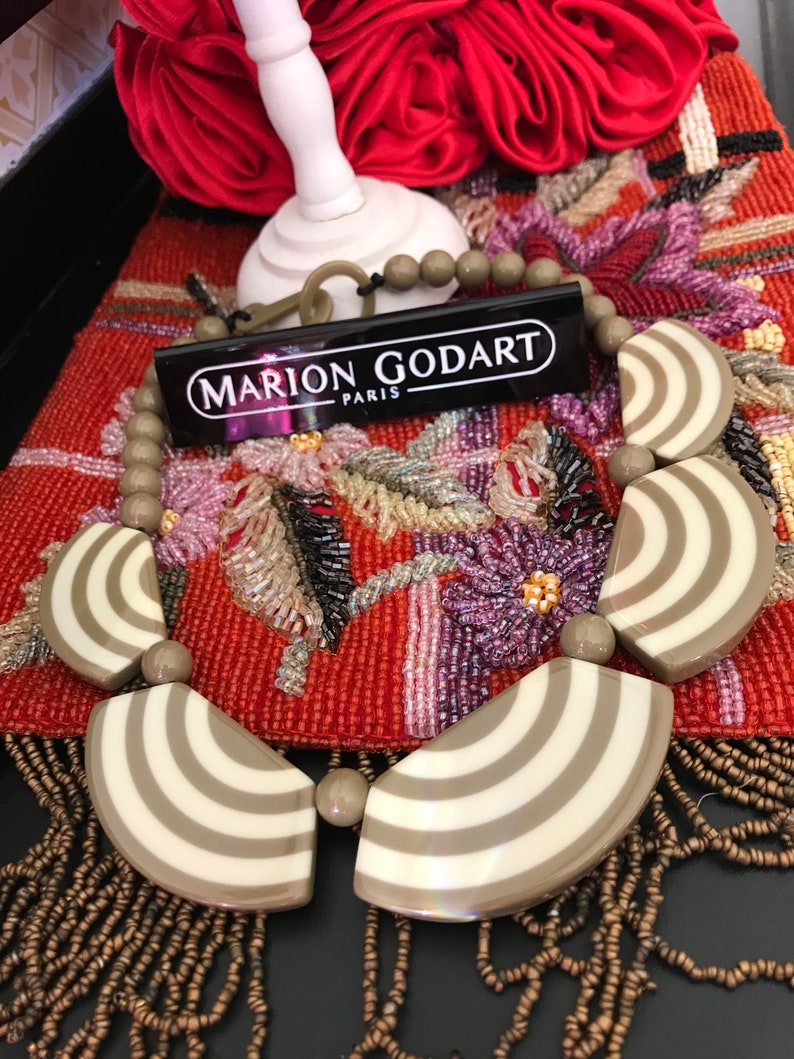May include: A striking necklace with large, fan-shaped pendants, cream and taupe striped design. The necklace has round, taupe beads and a tag reading "MARION GODART PARIS". It is displayed on a red, embroidered surface.