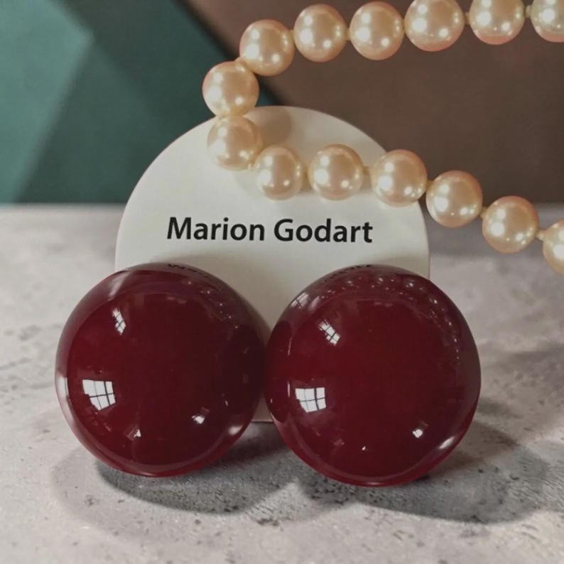 May include: A pair of round, deep burgundy earrings with a smooth, glossy finish. The earrings are displayed on a white circular card featuring the text Marion Godart in black lettering, positioned below a strand of cream-colored round pearls.