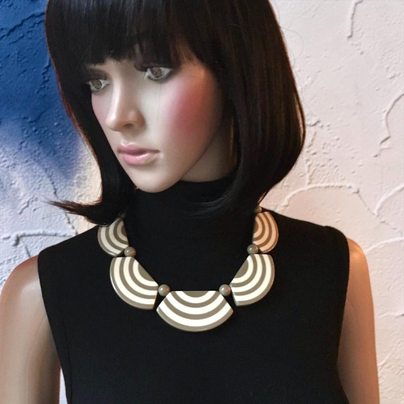 May include: A striking necklace with a geometric pattern of beige and white stripes. The necklace is made up of semi-circular shapes and small, round beads. The necklace is shown on a mannequin wearing a black turtleneck top.