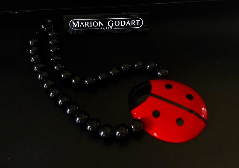 May include: A black beaded necklace with a red ladybug pendant. The ladybug has black spots and a black line down the center. The necklace is on a black surface. The tag reads "MARION GODART - PARIS".