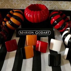 May include: A collection of Marion Godart Paris jewelry, including several chunky bracelets in red, black, orange, and white stripes. Also, square-shaped earrings in various colors are displayed on a black and white striped background.