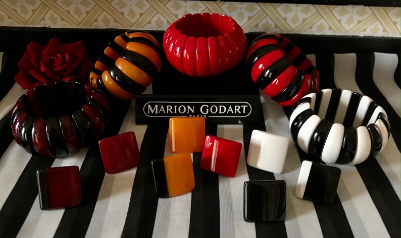 May include: A collection of Marion Godart Paris jewelry, including several chunky bracelets in red, black, orange, and white stripes. Also, square-shaped earrings in various colors are displayed on a black and white striped background.
