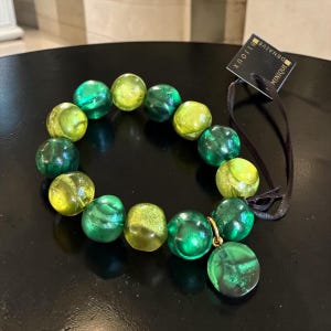 Peut inclure: Un bracelet élastique composé de perles en verre translucide rondes dans des tons vert citron et vert émeraude. Une petite breloque ronde en verre vert est suspendue à un anneau doré. Une étiquette noire porte le texte blanc "DOMINIQUE BIJOUX".