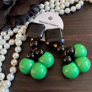 May include: A pair of dangle earrings with black and green beads. The earrings feature a black square-shaped bead at the top, followed by three green round beads and three black round beads.