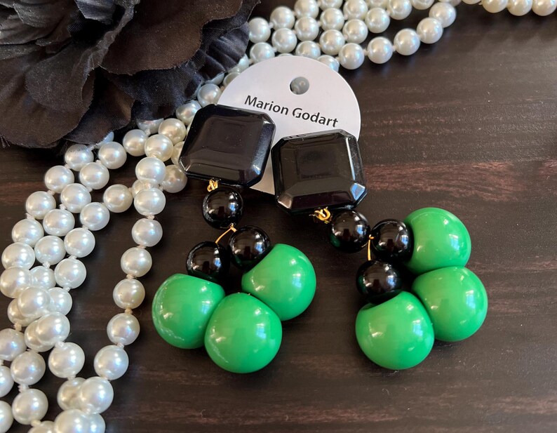 May include: A pair of dangle earrings with black and green beads. The earrings feature a black square-shaped bead at the top, followed by three green round beads and three black round beads.