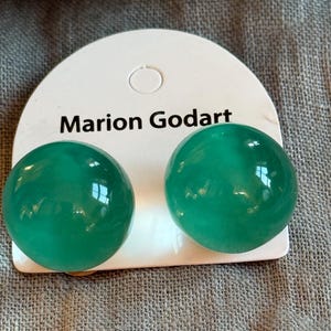 May include: A pair of round, emerald green earrings on a white Marion Godart branded card. The earrings are a glossy, spherical shape. The card is set against a textured, gray background.