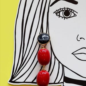 Puede incluir: Un par de pendientes colgantes con una parte superior negra y dorada y dos cuentas ovaladas rojas.