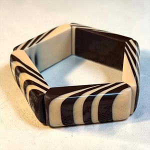 May include: A hexagonal bangle bracelet featuring a high-contrast pattern of alternating black and cream-colored stripes. The rigid, geometric accessory is constructed from smooth, polished segments that create a continuous, repeating wave-like design.