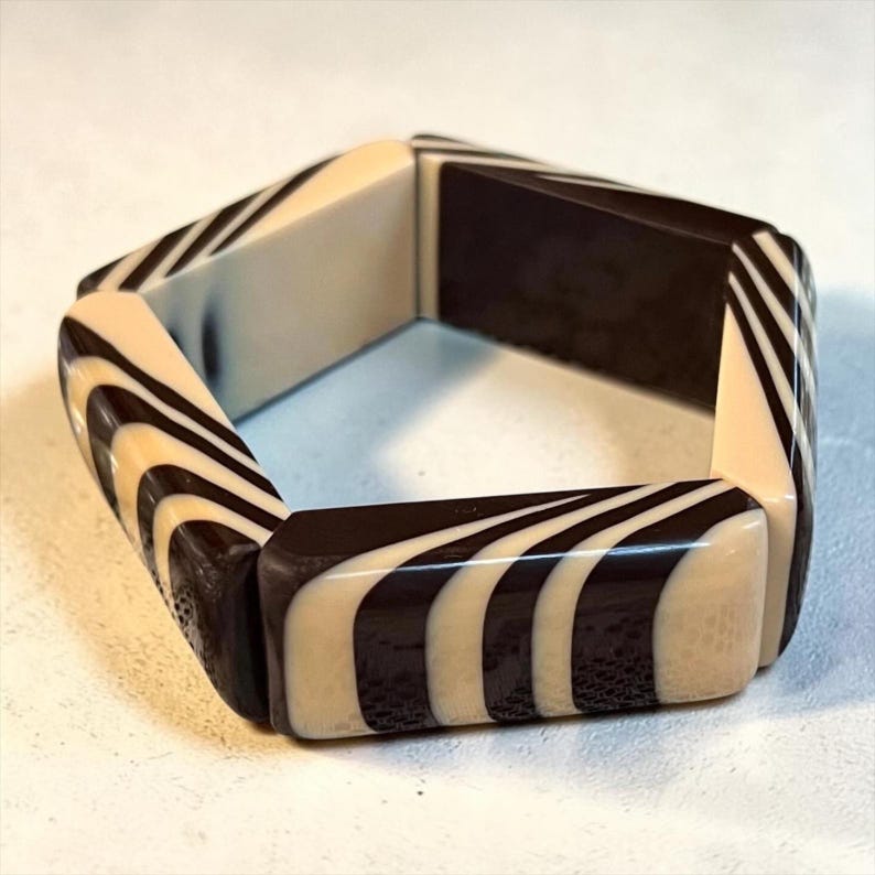 May include: A hexagonal bangle bracelet featuring a high-contrast pattern of alternating black and cream-colored stripes. The rigid, geometric accessory is constructed from smooth, polished segments that create a continuous, repeating wave-like design.