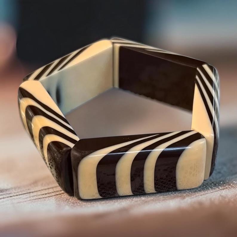 May include: A pentagonal bangle bracelet featuring a high-gloss finish with alternating segments of solid dark brown and cream-colored resin. The design displays a repeating pattern of curved, flowing stripes that wrap around each of the five flat sides.