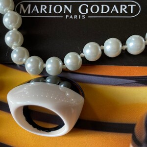 May include: A cream-colored ring with a black top and a pearl necklace. The ring has a rounded shape with a black interior. The necklace features large, round, off-white pearls. The text "MARION GODART PARIS" is visible in the background.