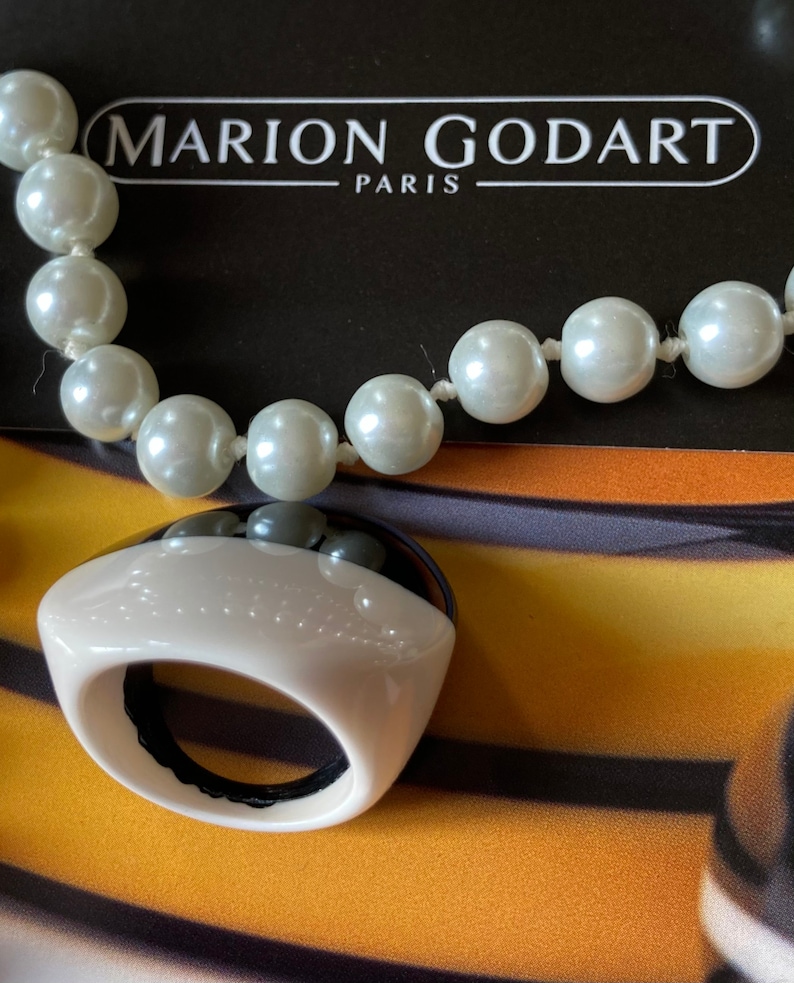 May include: A cream-colored ring with a black top and a pearl necklace. The ring has a rounded shape with a black interior. The necklace features large, round, off-white pearls. The text "MARION GODART PARIS" is visible in the background.
