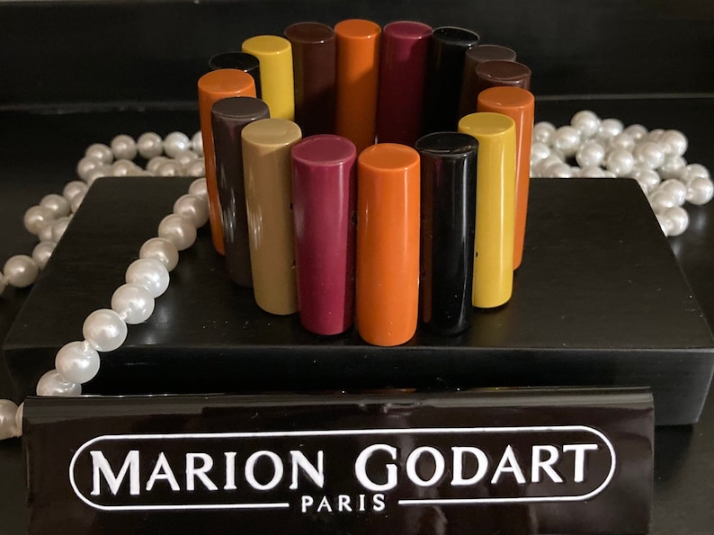 May include: A stretch bracelet composed of cylindrical resin segments in a multi-coloured palette of orange, yellow, brown, black, and magenta. The bracelet is displayed with a black rectangular plaque featuring the white text MARION GODART PARIS.