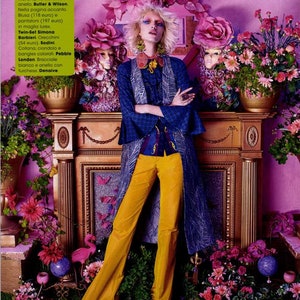 May include: A woman wearing a blue and white patterned jacket, yellow pants, and a necklace with a large flower pendant. She is standing in front of a fireplace with flowers and a pink flamingo figurine.