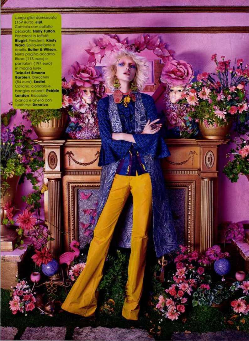 May include: A woman wearing a blue and white patterned jacket, yellow pants, and a necklace with a large flower pendant. She is standing in front of a fireplace with flowers and a pink flamingo figurine.