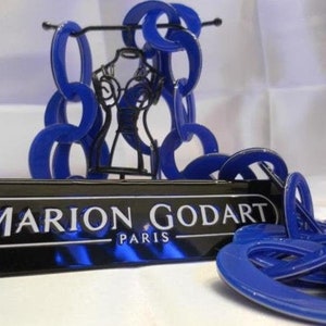 May include: A chunky blue resin chain necklace featuring large interlocking circular links. The necklace is displayed on a black wire mannequin stand behind a black rectangular plaque with the white text MARION GODART PARIS.
