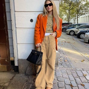 May include: An orange blazer, tan cargo trousers, and brown platform boots. A black handbag with a top handle is held. The outfit includes a patterned sweater and sunglasses. The setting is outdoors.