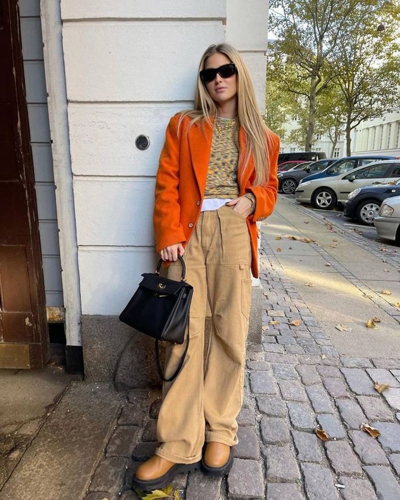 May include: An orange blazer, tan cargo trousers, and brown platform boots. A black handbag with a top handle is held. The outfit includes a patterned sweater and sunglasses. The setting is outdoors.