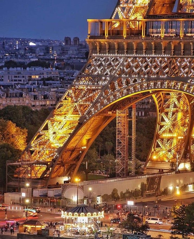 May include: A nighttime view of the Eiffel Tower in Paris, France. The tower is illuminated with golden lights, and the city lights can be seen in the background. A carousel is in the foreground, with people walking around it.