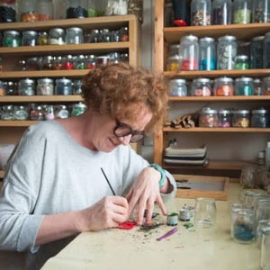 May include: A person with glasses is working on a craft project at a table. Jars filled with colorful materials are on shelves in the background. The person is using a paintbrush and small containers of paint.