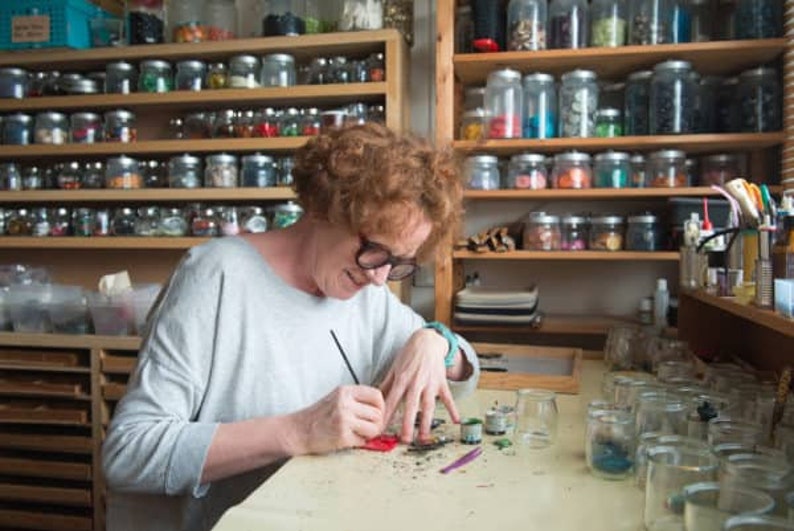 May include: A person with glasses is working on a craft project at a table. Jars filled with colorful materials are on shelves in the background. The person is using a paintbrush and small containers of paint.