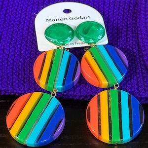 Könnte beinhalten: Bunte Marion Godart Ohrringe. Die Ohrringe haben einen grünen Stecker und einen großen, runden Anhänger mit Regenbogenstreifen. Die Ohrringe sind aus transparentem Material gefertigt und haben goldfarbene Beschläge. Sie werden auf einer lila Strickoberfläche präsentiert.