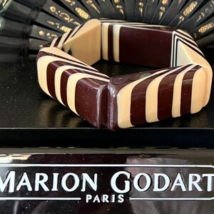 May include: A chunky bangle bracelet featuring a geometric pattern of alternating dark brown and cream-colored resin segments. The bracelet is displayed in front of a black plaque with the white text MARION GODART PARIS printed on the front surface.