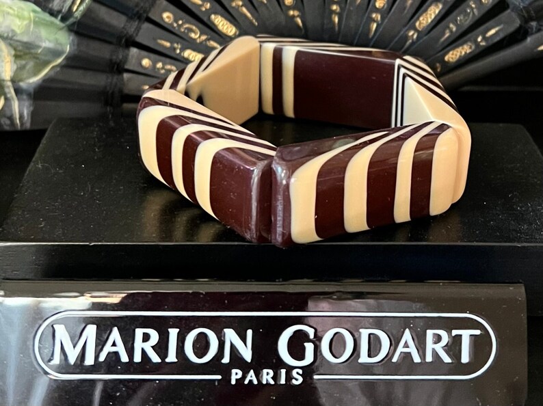 May include: A chunky bangle bracelet featuring a geometric pattern of alternating dark brown and cream-colored resin segments. The bracelet is displayed in front of a black plaque with the white text MARION GODART PARIS printed on the front surface.