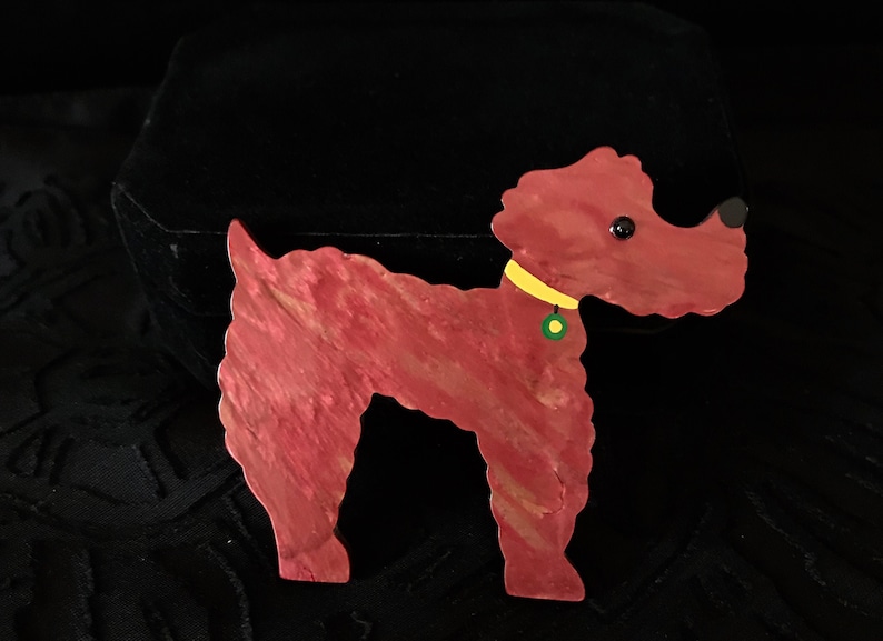 May include: A red and gold-toned dog-shaped brooch. The brooch has a yellow collar with a green and yellow tag. The dog has black eyes and a black nose. The brooch is made of a material with a marbled effect.