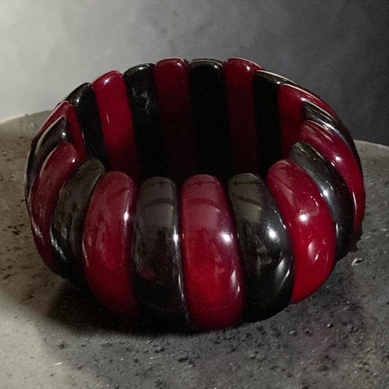 May include: A stretch bracelet composed of alternating smooth, rounded segments in deep glossy black and rich translucent dark red. The circular accessory features a repeating pattern of these polished, curved pieces arranged in a continuous band.