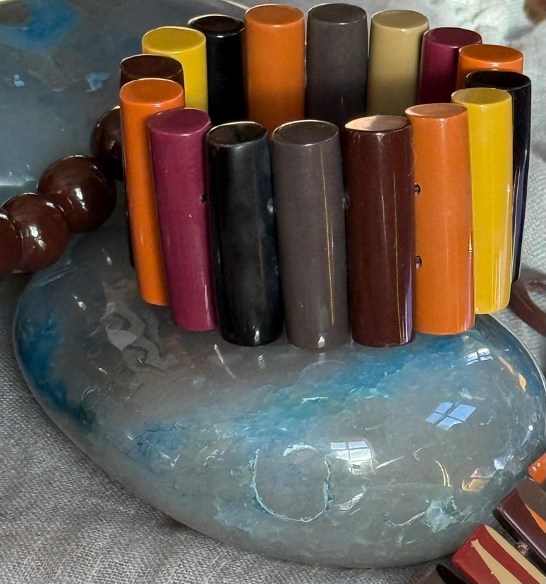 May include: A stretch bracelet composed of vertical, cylindrical beads in a variety of colours including orange, yellow, black, dark brown, maroon, and tan. The beads are arranged in a circular pattern, creating a multi-coloured, smooth, and glossy accessory.