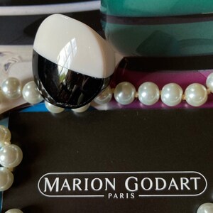 May include: A black and white oval-shaped object with a white top, resting on a black surface. The object is next to a strand of white pearls and a black card with the text "MARION GODART PARIS". A green and purple decorative object is in the background.