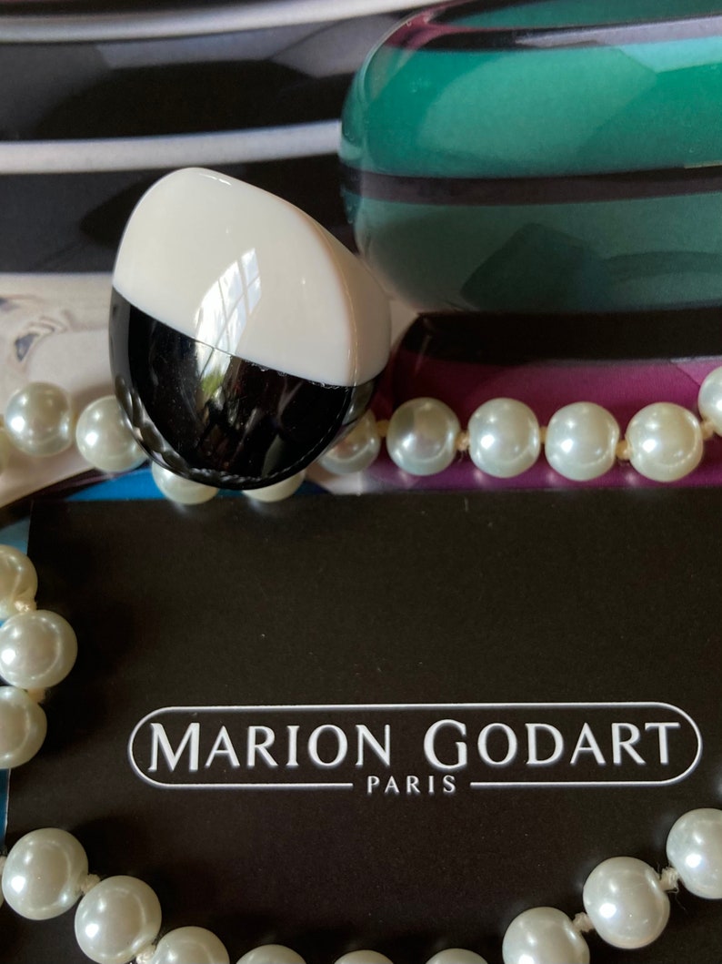 May include: A black and white oval-shaped object with a white top, resting on a black surface. The object is next to a strand of white pearls and a black card with the text "MARION GODART PARIS". A green and purple decorative object is in the background.