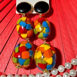 Puede incluir: Un par de aretes largos y coloridos con una parte superior negra y dorada. Los aretes tienen dos dijes ovalados llenos de formas geométricas coloridas. Los dijes están conectados por una cadena dorada. Los aretes se muestran sobre un fondo rojo con una etiqueta blanca que dice "Marion Godart".