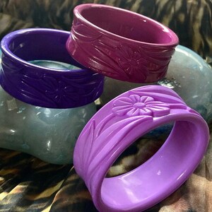 May include: Three purple plastic bangles with floral designs. The bangles are stacked on top of each other.