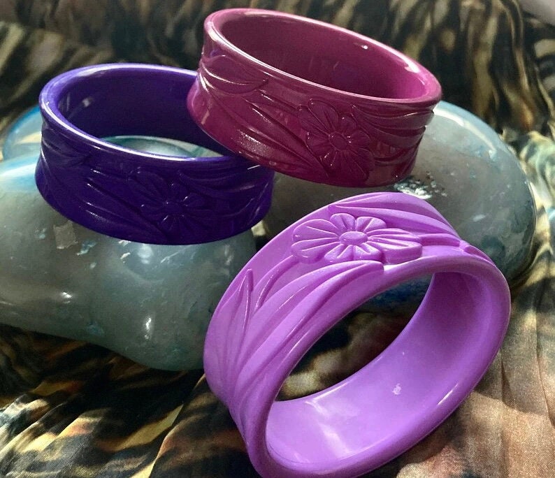 May include: Three purple plastic bangles with floral designs. The bangles are stacked on top of each other.