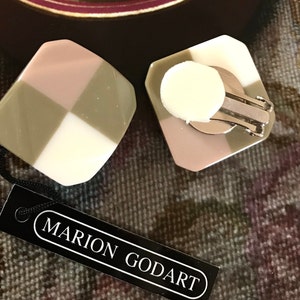 May include: Geometric clip-on earrings with a checkerboard design in white, pink, and olive green. The earrings are branded Marion Godart, as shown on the black tag.