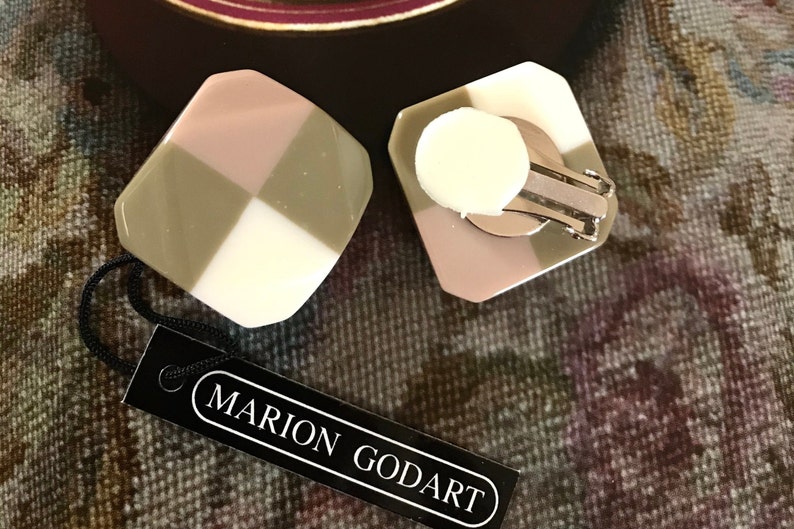 May include: Geometric clip-on earrings with a checkerboard design in white, pink, and olive green. The earrings are branded Marion Godart, as shown on the black tag.