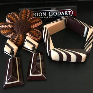 May include: A Marion Godart Paris jewelry set featuring a pentagonal bangle and two drop earrings with a dark brown and cream striped pattern. A brown and tan crocheted flower accessory is positioned behind the earrings and the bangle.