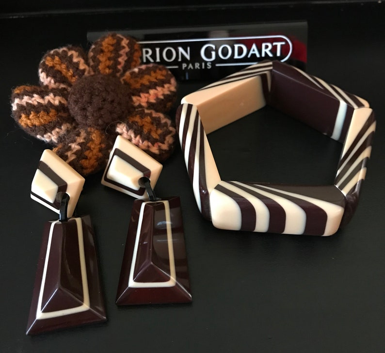 May include: A Marion Godart Paris jewelry set featuring a pentagonal bangle and two drop earrings with a dark brown and cream striped pattern. A brown and tan crocheted flower accessory is positioned behind the earrings and the bangle.