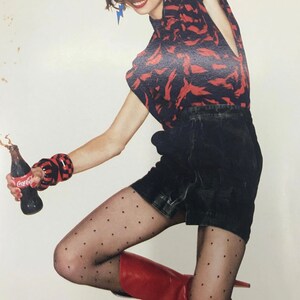 May include: A woman wearing a red and black patterned shirt, black denim shorts, and black polka dot tights. She is holding a bottle of Coca-Cola.