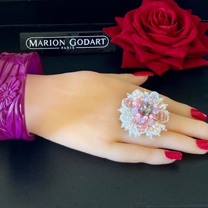 May include: A purple plastic bangle bracelet with a floral design. The bracelet is on a mannequin hand wearing a large white ring with pink and silver embellishments.