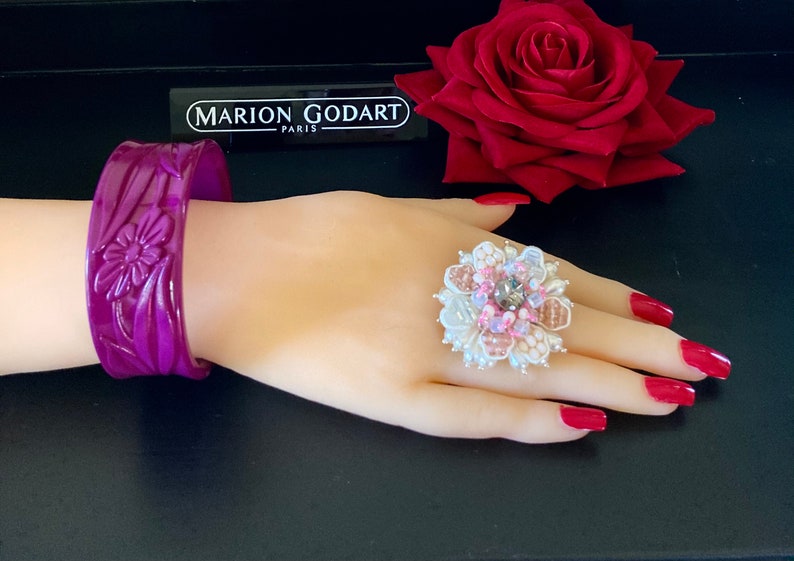 May include: A purple plastic bangle bracelet with a floral design. The bracelet is on a mannequin hand wearing a large white ring with pink and silver embellishments.